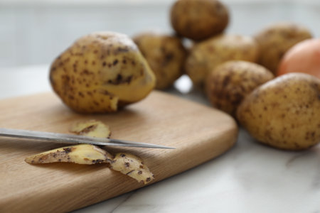 Fresh potatoes, peels, wooden board and knife on white marble table, closeupの写真素材