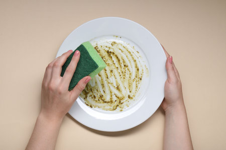 Woman washing dirty plate with sponge on beige background, top view
