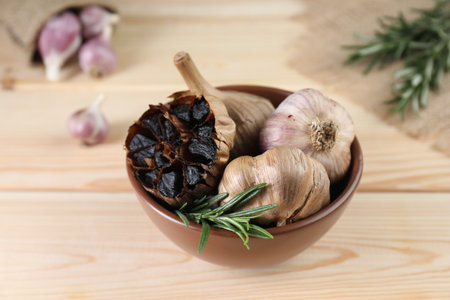 Bulbs of fermented black garlic and rosemary in bowl on wooden tableの写真素材