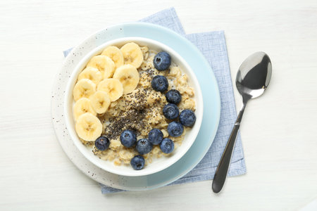 Tasty oatmeal with banana, blueberries and chia seeds served in bowl on white wooden table, top viewの写真素材