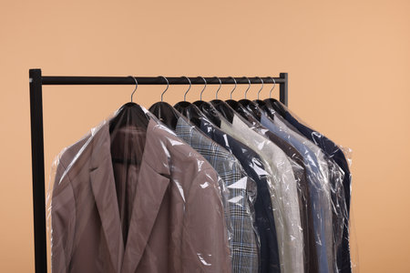 Dry-cleaning service. Many different clothes in plastic bags hanging on rack against beige backgroundの写真素材