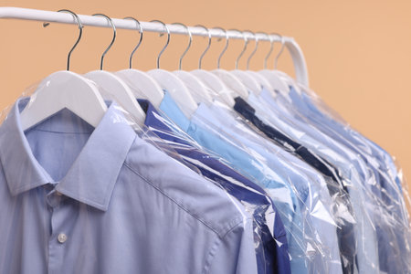 Dry-cleaning service. Many different clothes in plastic bags hanging on rack against beige background, closeupの写真素材