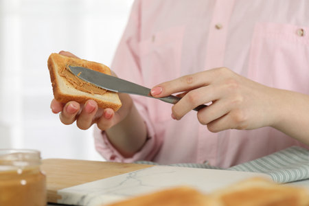 Woman spreading tasty nut butter onto toast at table, closeup