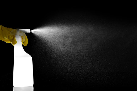 Woman spraying liquid from bottle on black background, closeup