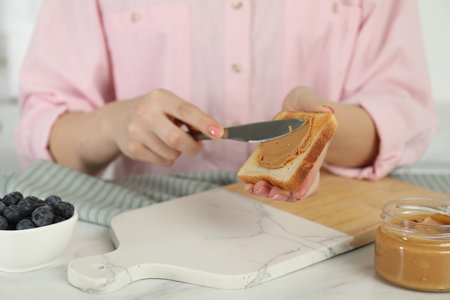 Woman spreading tasty nut butter onto toast at white table, closeup