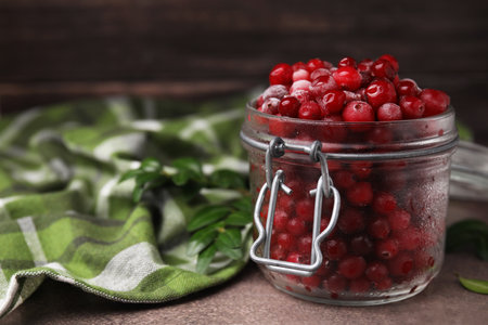 Frozen red cranberries in glass jar and green leaves on brown textured table, closeup. Space for textの写真素材