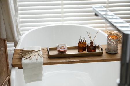 Wooden tray with cosmetic products, burning candles, reed air freshener and towel on bath tub in bathroomの写真素材