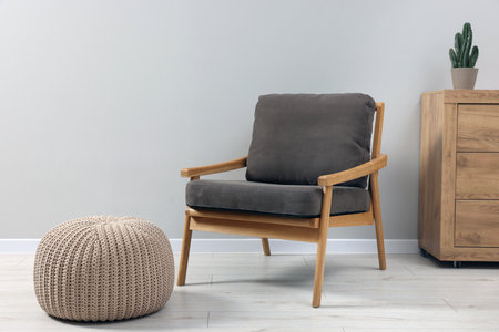 Comfortable armchair, chest of drawers and pouf near light gray wall indoors. Interior design