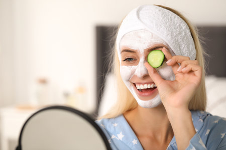 Young woman with face mask and cucumber looking into mirror at home, space for text. Spa treatmentsの写真素材