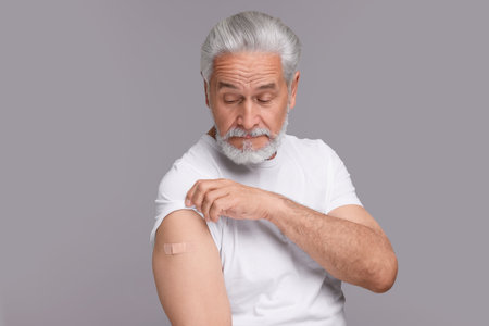 Senior man with adhesive bandage on his arm after vaccination against light gray background
