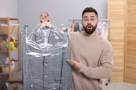 Dry-cleaning service. Shocked man holding hanger with jacket in plastic bag indoorsの写真素材