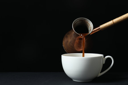 Turkish coffee Pouring brewed beverage from cezve into cup on table against black background, space for textの写真素材