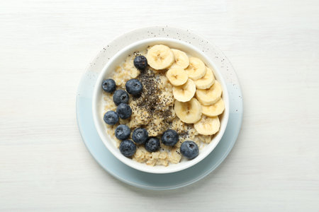 Tasty oatmeal with banana, blueberries and chia seeds served in bowl on white wooden table, top viewの写真素材