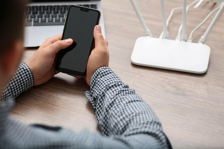 Man with smartphone and laptop connecting to internet via Wi-Fi router at wooden table, closeup