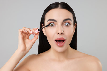 Emotional young woman applying mascara on gray backgroundの写真素材