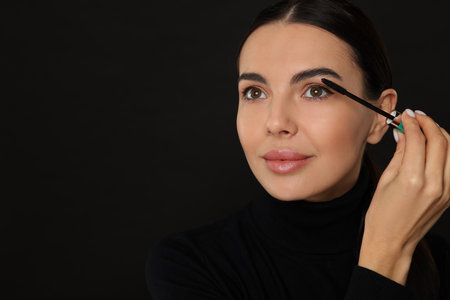 Beautiful young woman applying mascara on black background, closeup. Space for textの写真素材