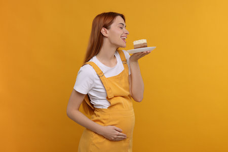 Pregnant young woman eating piece of tasty cake on orange backgroundの写真素材