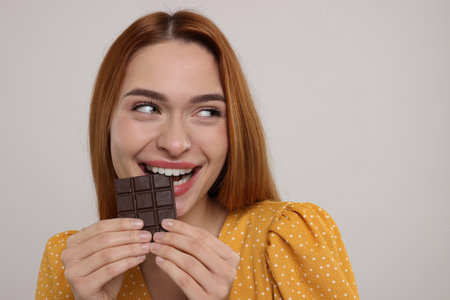 Young woman eating tasty chocolate on light gray background, space for text