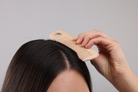 Woman with comb examining her hair and scalp on gray background, closeup