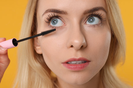 Beautiful woman applying mascara on orange background, closeupの写真素材