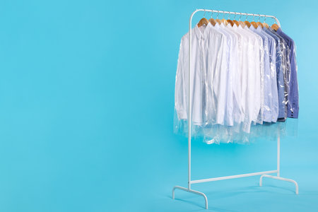 Dry-cleaning service. Many different clothes in plastic bags hanging on rack against light blue background, space for textの写真素材