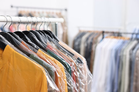 Dry-cleaning service. Many different clothes in plastic bags hanging on rack indoors, closeup