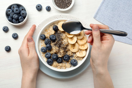 Woman eating tasty oatmeal with banana, blueberries and chia seeds at white wooden table, top viewの写真素材