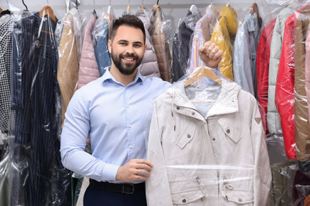 Dry-cleaning service. Happy worker holding hanger with jacket in plastic bag near other clothes indoorsの写真素材