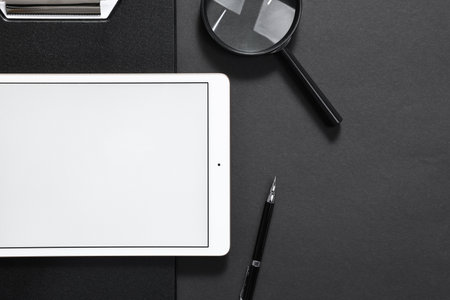 Modern tablet, clipboard, pen and magnifying glass on black background, flat lay. Space for text