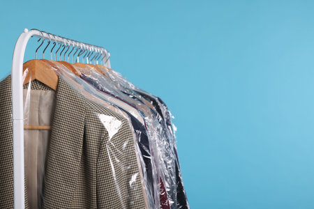 Dry-cleaning service. Many different clothes in plastic bags hanging on rack against light blue background, space for textの写真素材