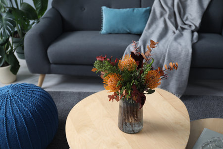 Vase with bouquet of beautiful leucospermum flowers on wooden nesting tables, sofa and ottoman indoorsの写真素材
