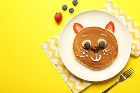 Creative serving for kids. Plate with cute cat made of pancakes, berries, cream, banana and chocolate paste on yellow background, flat lay. Space for textの写真素材