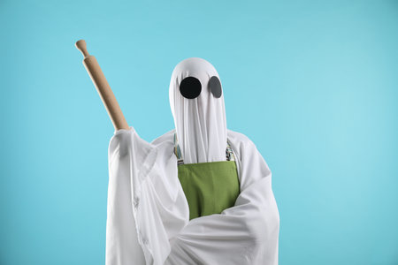 Woman in ghost costume and apron with rolling pin on light blue backgroundの写真素材