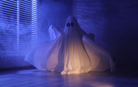 Creepy ghost. Woman covered with sheet near window in blue lightの写真素材