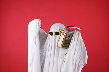 Person in ghost costume and sunglasses using retro radio receiver on red backgroundの写真素材