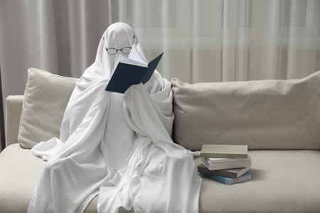 Creepy ghost. Person covered with white sheet reading book on sofa at homeの写真素材