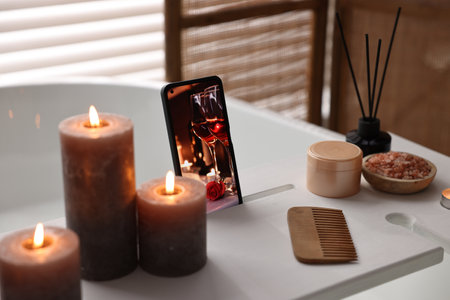 White wooden tray with smartphone, burning candles and beauty products on bathtub in bathroom