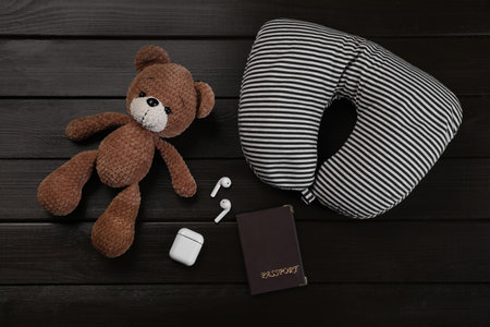 Striped travel pillow, toy bear, passport and earphones on dark wooden background, flat lay