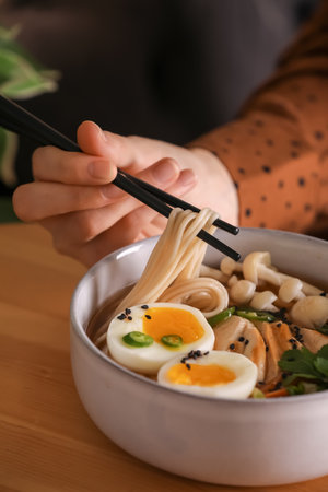 Woman eating delicious ramen with chopsticks at wooden table, closeup. Noodle soupの写真素材