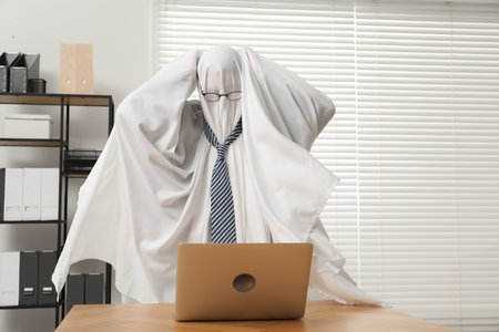 Overworked ghost. Man covered with white sheet using laptop at wooden table in officeの写真素材