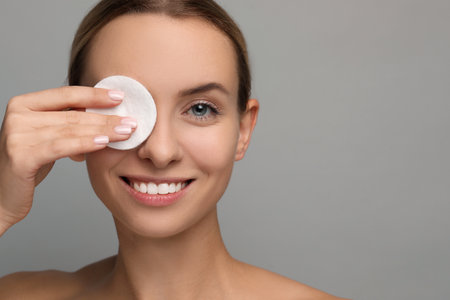 Smiling woman removing makeup with cotton pad on gray background, closeup. Space for textの写真素材