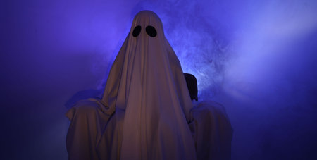 Creepy ghost. Woman covered with sheet sitting in armchair in blue lightの写真素材
