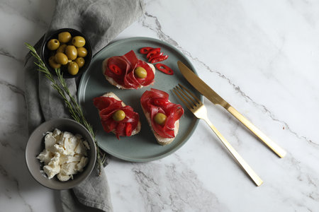 Delicious sandwiches with bresaola, cream cheese and olives served on white marble table, flat lay....