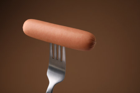 Delicious boiled sausage on fork against brown background, closeupの写真素材