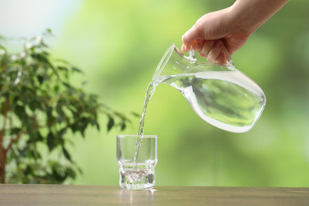 Woman pouring fresh water from jug into glass at wooden table against blurred green background, closeup. Space for textの写真素材