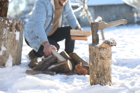 Man taking logs from pile outdoors on winter day. Metal ax in wooden log on snow, selective focusの写真素材