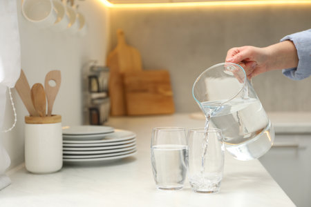 Woman pouring water from jug into glass at white table in kitchen, closeup. Space for textの写真素材