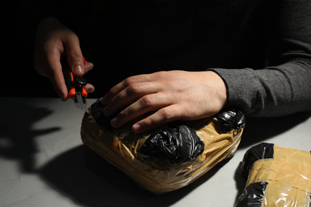 Smuggling and drug trafficking. Man opening package of narcotics with box cutter in darkness,...