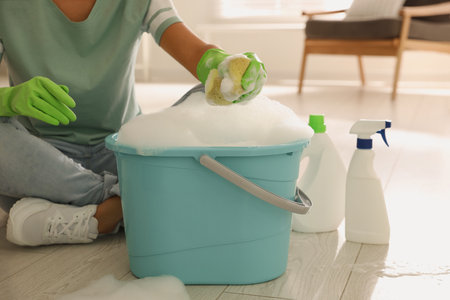 Woman holding sponge with foam over bucket indoors, closeup. Cleaning suppliesの写真素材