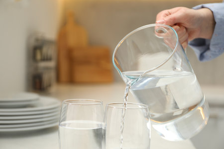 Woman pouring water from jug into glass at white table in kitchen, closeup